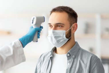 Doktor, evdeki gençlerin sıcaklığını elektronik termometreyle ölçüyor. Beyaz maskeli adam, koronavirüs aşısından önce sağlığını kontrol ediyor. Covid konseptine karşı aşı