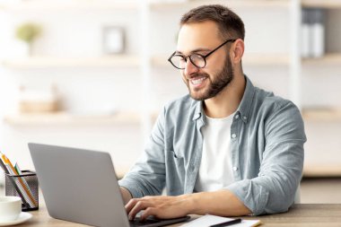 ABD bayrağı taşıyan mutlu genç adam ofis laptopuyla masa başında çalışıyor. Yabancı iş, eğitim veya Amerika 'ya göç için İngilizce öğrenen milenyum erkek öğrencisi
