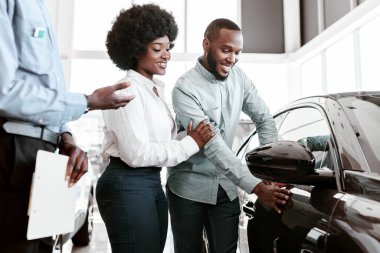 Araba satış işi. Yönetici Afro çiftle konuşuyor, galerideki yeni otomobili gösteriyor. Genç siyahi aile araç seçiyor, galeride modern otomobillere bakıyor.
