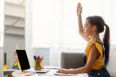 Evde eğitim kavramı. Profil yan görüntüsü: Kanepede oturan odaklanmış öğrenci, video görüşmesi sırasında cevap için elini kaldırıyor ve öğretmeninin sanal dersi var, boş ekran.