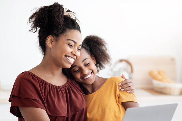 Симпатичные чернокожие мать и ребенок вместе используют ноутбук, сидят за кухонным столом с домашней выпечкой, обнимаются и смотрят на экран компьютера, проводят время вместе, смотрят мультики, копируют пространство
