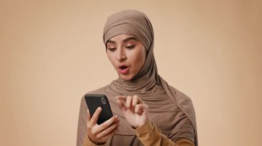 Bej tesettürlü genç bir Arap kadın, akıllı telefonuyla nötr bir arka planda etkileşim kurarken gülümsüyor. Günlük hayatta modern iletişim ve pozitiflik sergiliyor..