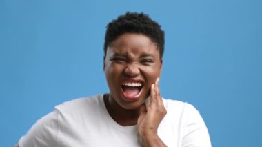 Genç bir Afrikalı Amerikalı kadın mavi arka plan karşısında duruyor, yanağını tutarak ve acı içinde yüzünü buruşturarak rahatsızlığını gösteriyor. Yüzündeki ifade bir sıkıntı anını yansıtıyor..