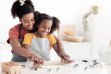 Neşeli Afro-Amerikan anne ve kızı ev yapımı hamur işleri ve gülümseme için hamuru yuvarlıyor, evde birlikte yemek pişirirken eğleniyorlar, önlük giyiyorlar, mutfak önlüğü giyiyorlar, fotokopi çekiyorlar.