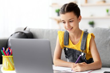 Akademik kavram. Masasında oturmuş, dizüstü bilgisayar kullanarak kağıt defterine renkli kalemle bakarak ya da çizerek gülümseyen genç kız. Webinar izlerken not alan liseli kız