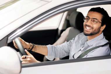 Bin yıllık mutlu orta doğulu adam güzel bir araba kullanıyor, gözlük takıyor, yandan bakıyor, dışarıdan çekiliyor, fotokopi çekiyor. Pozitif Hintli adam direksiyonda el ele tutuşup yola bakıyor ve gülümsüyor.