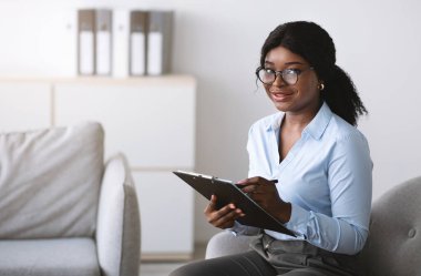 Konsültasyon sırasında not alan mutlu siyah kadın psikolog, zihinsel sağlık kliniğinde kameraya gülümsüyor, boş alan. Ofisteki koltukta oturan genç psikoterapist.