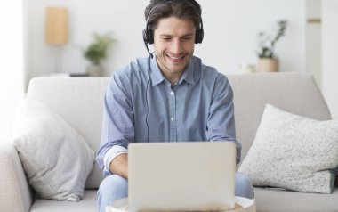 Kulaklıklı Pozitif Adam dizüstü bilgisayar kullanıyor Podcast dinliyor ya da evde koltukta oturan Webinar 'da rol alıyor.