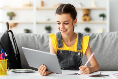 Çocuk ve Eğitim Konsepti. Not defterinde ödev yapan genç kızların portresi masadaki koltukta oturuyor, tablet bilgisayarını tutuyor ve kullanıyor, ev eğitiminden keyif alıyor.