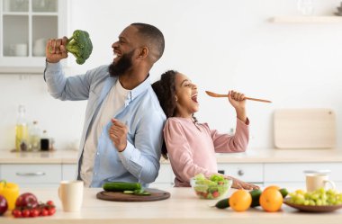 Mutfak eğlencesi. Pozitif Afrikalı Amerikalı baba ve küçük kızının portresi mutfakta birlikte yemek pişirirken şarkı söyleyip dans etmeleri, spatula ve brokoliyi mikrofon olarak kullanmaları, yemek hazırlamaları.