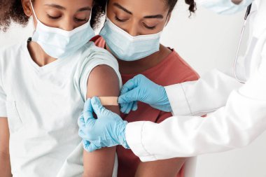 Afro-Amerikan ailesi genç anne ve genç kız klinikte aşı yapıyorlar, tıbbi maske takıyorlar. Siyahi kız için COVID-19 iğnesinden sonra hemşire omzuna bant takıyor.