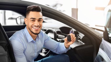 Yakışıklı Arap müşteri arabada oturuyor ve anahtarı tutuyor, kameraya gülümsüyor, galeriden araba alıyor. Neşeli orta doğulu adam güzel bir spor arabanın içinde oturuyor, yeni bir araba aldığına seviniyor.