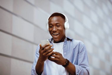 Heyecanlı kulaklıklı Afrikalı Amerikalı adam tuğla duvarın yanındaki akıllı telefon ekranına bakıyor.