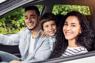 Cheerful glad millennial arab mother and father with kid go on vacation by auto and look at camera at open window. Family trip on new car, weekend together, love and relationship, positive emotions