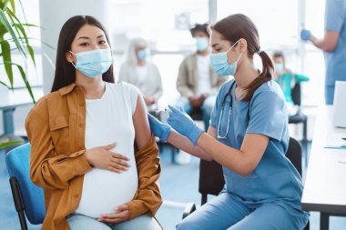 Hamilelik Aşısı. Hamile Asyalı kadın grip önleme aşısı oluyor, hemşire Coronavirüs 'e aşı yapıyor, anne adayı kliniğe enjekte ediyor, maske takıyor ve eldiven takıyor.