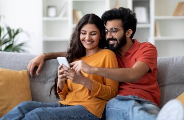 Mutlu Hintli çiftin portresi, akıllı telefonuyla koltukta dinlenirken gülümseyen Doğu eşleri internetten alışveriş yaparken ya da oturma odasında dinlenirken yeni mobil uygulamalara bakarken,