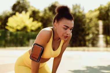 Aktif eğitim. Sarı spor giyim sutyen ve kol bandı takmış yorgun Afrikalı Amerikalı genç bir kadın. Sabah koşusu sırasında hücrede dinleniyor. Açık havada, parkta ya da spor alanında soluklanıyor.