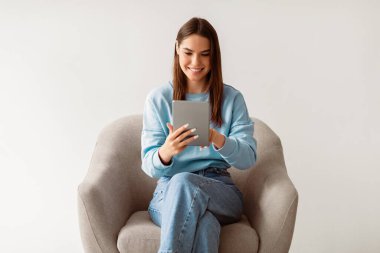 Beyaz kadın dijital tablet, internet ya da sosyal medya kullanıyor, koltukta oturuyor, modern teknolojilerin tadını çıkarıyor, beyaz stüdyo arka planında yeni uygulama ya da web sitesini kontrol ediyor.