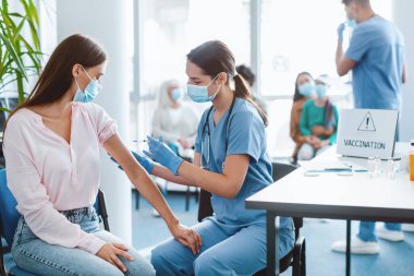 Grip aşısı kampanyası. Koluna Coronavirüs aşısı yapılan genç bir bayan. Hastanede doktor ya da hemşireyle oturuyor. Corona Virüsü Bağışıklık Sistemi, Tedavi ve Tedavi