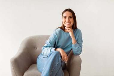 Mutlu bayan. Sıradan kıyafetler giyen, kameraya bakan, koltukta oturan, beyaz stüdyo arka planında poz veren, güler yüzlü beyaz bir kadın. Gerçek insanların portreleri.