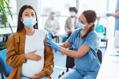 Hamile Asyalı Kadın gribe karşı aşı oluyor, Sağlık Merkezi 'nde oturuyor, Karnına dokunuyor, Coronavirus antiviral aşı enjekte ettikten sonra alçı uygulayan kadın