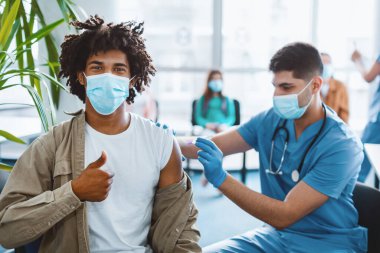 Aşı ol. Gülümseyen Afro-Amerikalı genç adam klinikteki koronavirüs aşısı sırasında baş parmak hareketi gösteriyor, covid-19 aşısını teşvik ediyor, iğneleniyor.