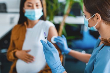 Küresel Salgın sırasında grip aşısı. Hastanede yüz maskesi takmış hamile bir kadın iğne oluyor. Doktor şırıngayı iğne yapmaya hazırlıyor. Kolunu pamukla ovuyor, iğneye odaklanıyor.