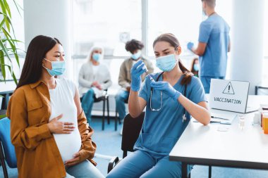 Tıbbi maske takan genç bir kadın doktor ya da hemşire aşı kampanyası sırasında hamile Asya 'lı kadına grip aşısı enjekte etmeden önce ilaç hazırlıyordu.