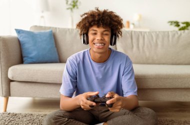 Evde kulaklık takan, çevrimiçi video oyunları oynayan havalı siyahi genç oyuncu portresi. Afrika kökenli Amerikalı ergenler kapalı alanda karantina sırasında eğlenceli bir hobi ediniyorlar.