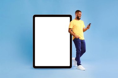 İyimser genç Afrikalı Amerikalı adam, beyaz ekranlı büyük tablet bilgisayara yaslanıyor, cep telefonunu mavi stüdyo arka planında kullanıyor, web sitesi veya uygulama tasarımı için model alıyor.