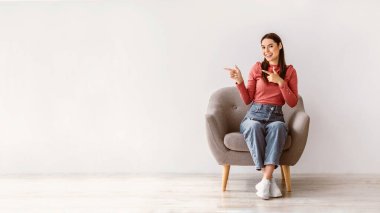 Güzel teklif. Koltukta oturan mutlu genç bayan, beyaz stüdyo duvarına karşı boş alanı işaret ediyor, pankart tasarımı. Reklam ya da tanıtım metinleri sunan olumlu milenyum kadını