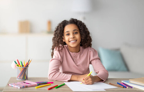 Счастливая чернокожая школьница рисует картину с ручками и маркерами, сидит за партой и улыбается фотоаппарату. Творческая школьница наслаждается своим новым хобби, домашним развлечением