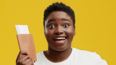 Tombul vücutlu genç bir Afrikalı Amerikalı kadın elinde parlak sarı bir arka plana karşı bir pasaport tutarken geniş bir gülümseme sergiliyor. Heyecan ve neşe gösteriyor..