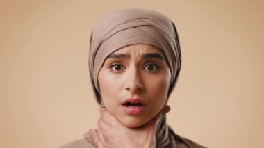 Türbanlı genç bir Arap kadın, bir stüdyoda tarafsız bir arkaplanda boynunu tutarken şaşırmış bir ifade sergiliyor. Bu çarpıcı görüntü yoğun bir duygu anını yakalar..