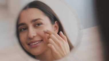 Rahat bir ev ortamında, genç bir Orta Doğulu kadın yansımasına gülümserken nazikçe cilt ürünlerini uyguluyor. Güzelliğe odaklanmış barışçıl bir kişisel bakım rutini yakaladığımız an.