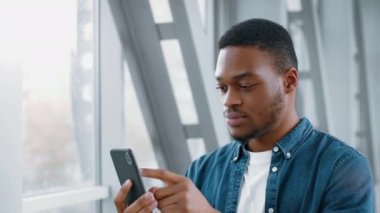 Sıradan giyinmiş genç bir Afrikalı Amerikalı havaalanında beklerken akıllı telefonuna odaklanıyor. Cihazıyla yönünü değiştirirken meşgul ve dikkatli görünüyor..