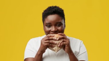 Tombul genç bir kadın heyecanla stüdyo ortamında büyük bir burger yemeye hazırlanıyor. Parlak sarı arka plan, yemeğe odaklanmış neşeli ifadesini vurguluyor..