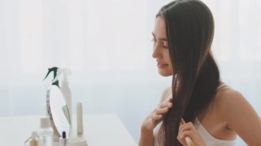Genç bir Orta Doğulu kadın parlak bir odada kendini şımartıyor. O, aynadaki yansımasını hayranlıkla izlerken ürünlerini kullanarak saç bakım rutinine odaklanmış durumda..