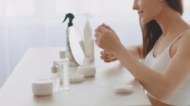 Genç bir Ortadoğulu kadın aynaya bakıp cilt rutinine odaklanarak serum uyguluyor. O çeşitli güzellik ürünleriyle dolu güneşli bir odada..