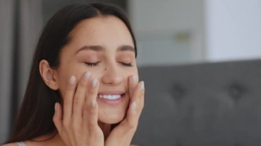 Genç bir Arap kadın evde cildini şımartırken kapalı gözlerle gülümsüyor. Günlük güzellik rutininin bir parçası olarak yüz kremi sürüyor. Kendine özen ve rahatlığı kucaklıyor..
