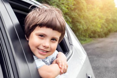 Gülümseyen küçük çocuk arabaya biniyor, açık pencereden dışarı bakıyor, eğleniyor, araba yolculuğunun tadını çıkarıyor, yazın açık havada, kapalı, boş alan. Aileyle hafta sonu gezisi, yol ve tatil, otomobil yolculuğu.