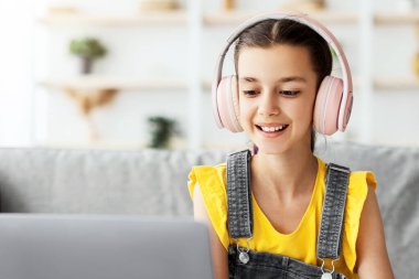 Uzaktan eğitim konsepti. Gülümseyen genç kız kanepede oturuyor, dizüstü bilgisayar kullanıyor ve not defterine yazıyor. Kulaklıklı kız öğrenci çevrimiçi ders izliyor, notlar alıyor, ödev yapıyor.