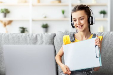 Gülümseyen kız öğretmeniyle video görüşmesi yapıyor, elinde kağıt tutarak ödevin sonucunu dizüstü bilgisayarla gösteriyor. Uzaktan çalışan bir çocuk. Yaratıcılık dersleri, ev okulu