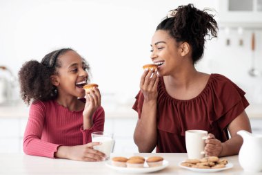 Güzel siyah anne ve genç kız kurabiye yiyorlar, mutlu Afrikalı Amerikalı kız sevgili annesiyle kahvaltı ediyor, mutfak içi. Sağlıklı beslenme, kalsiyum, çocuk gıda konseptinde vitamin.