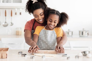 Mutlu siyah anne, ergen çocuğuna kurabiye pişirmeyi öğretiyor, neşeli Afro-Amerikan anne ve kızı ev yapımı hamur işleri için rulo yapıyor ve gülümsüyor, önlük giyiyor, mutfak önlüğü giyiyor.