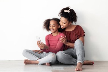 Güzel siyah anne ve kız birlikte cep telefonu kullanıyor, yerde oturuyor, sosyal medya için selfie çekiyor, evde birlikte vakit geçiriyorlar, beyaz boş duvar arka planı, kopyalama alanı.