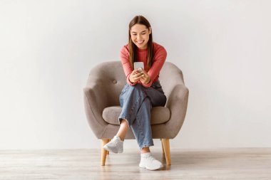 Mutlu beyaz kadın cep telefonu kullanıyor, internette sohbet ediyor, çalışıyor ya da internetten çalışıyor, beyaz stüdyo duvarına karşı koltukta oturuyor. Neşeli genç bayan cep telefonundan video izliyor.