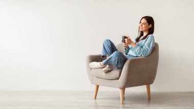 Neşeli genç bir kadın, beyaz duvara karşı rahat koltukta rahatça otururken aromatik kahve içerken, boş alanı olan bir panorama. Gülümseyen milenyum bayanı sıcak bir içecek içiyor, molasında dinleniyor.