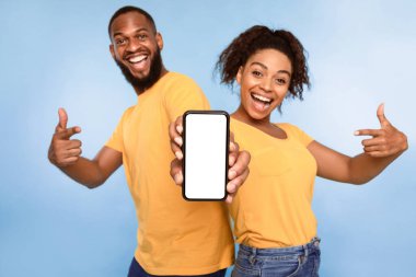 Mobil uygulama reklamı. Heyecanlı Afro-Amerikalı çift cep telefonlarını boş ekranla işaret ediyor, mavi stüdyo arka planında yeni bir uygulama öneriyorlar, seçici odaklanma. Web sitesi için model