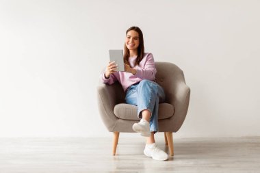 Modern teknoloji ve iletişim. Tabletli neşeli genç bir kadın koltukta oturuyor, çalışıyor ya da internetten çalışıyor, beyaz stüdyo duvarına karşı görüntülü sohbet yapıyor.